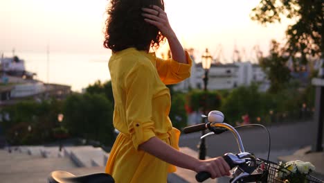 attractive brunette woman in a long yellow dress enjoying the dawn standing by her city bicycle holding its handlebar with