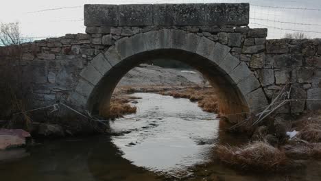 Vuelo-Lento-Y-Muy-Bajo-Bajo-Un-Pequeño-Puente-De-La-época-Romana-En-Invierno,-Muy-Cerca-De-La-Superficie-Del-Río-Que-Pasa-Por-Debajo-Del-Puente