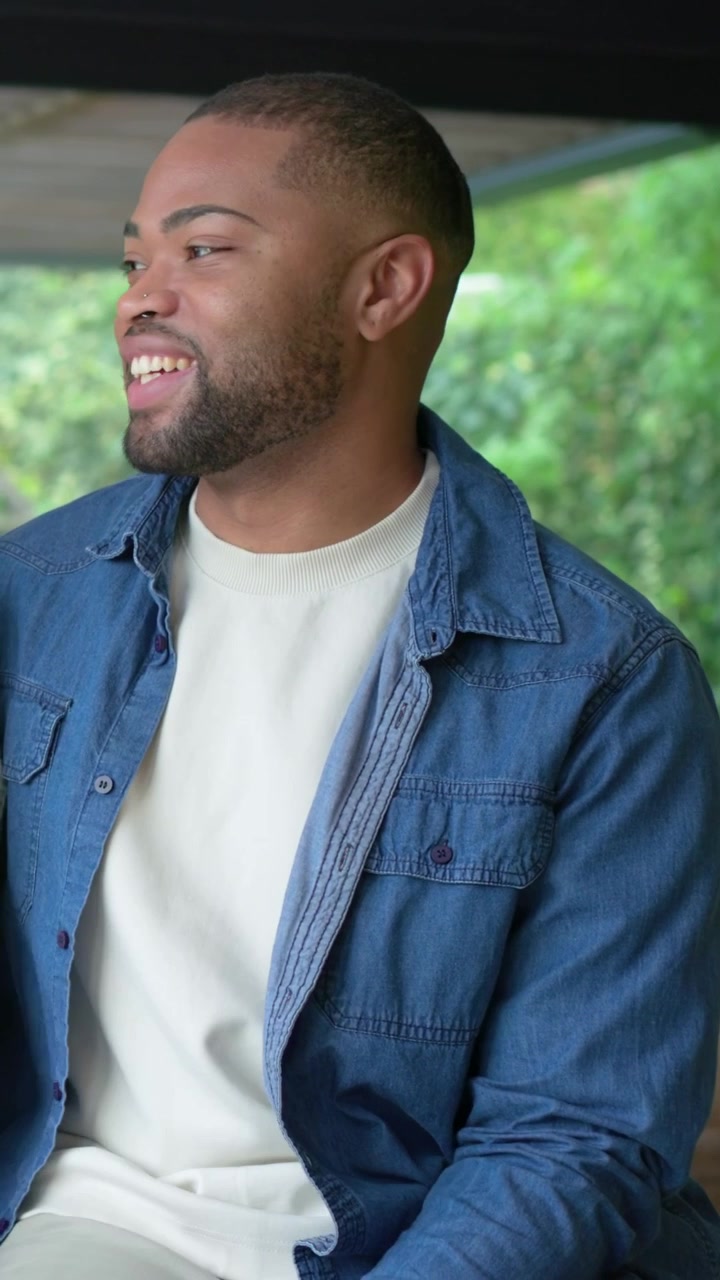 Vertical video: Turning to companion after companion speaking, man in denim shirt smiling on porch