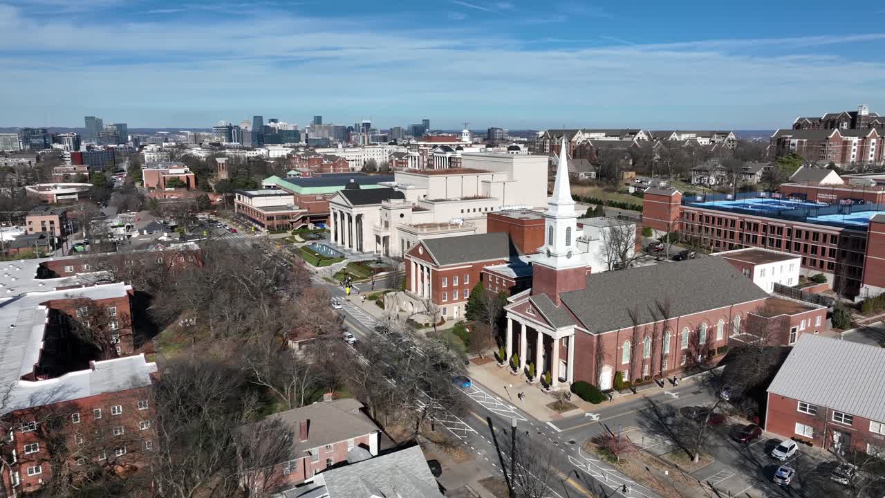 Vista aerea ascendente della Belmont University a Nashville, Tennessee, che mostra edifici del campus in mattoni rossi e il campanile della chiesa sotto un cielo azzurro limpido con lo skyline del centro città in lontananza
