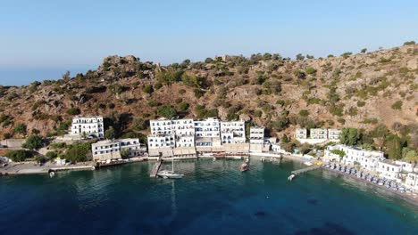 Drone-view-in-Greece-flying-over-blue-sea-in-Loutro-small-white-house-town-and-small-boats-next-to-a-hill-on-a-sunny-day