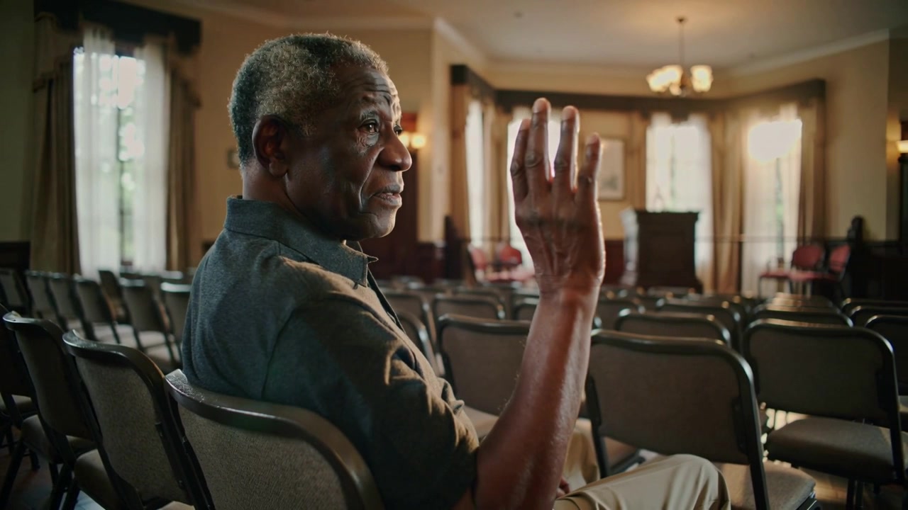 Responding to off-frame prompt, senior man in dark polo raising right hand in chapel chairs