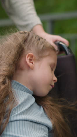 little girl with cerebral palsy sleeps in wheelchair at walk with mommy in city park. woman takes care of daughter spending time on fresh air closeup