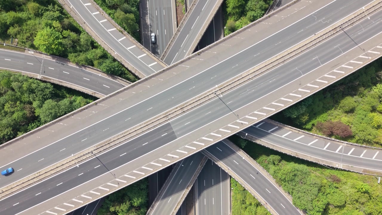 An aerial view showcases a multi-lane highway interchange surrounded by green trees. Cars, vans, and a large truck travel smoothly on the various roads and ramps during clear daytime conditions.