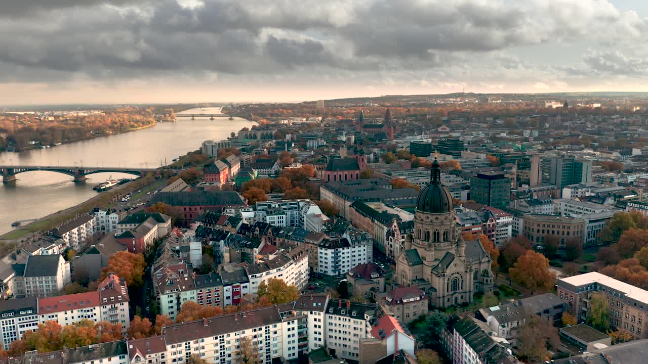 Weite Drohnenaufnahme der Christuskirche Mainz im Herbst mit goldenem Licht im Hintergrund und braunen Blättern am Baum aus der Luft