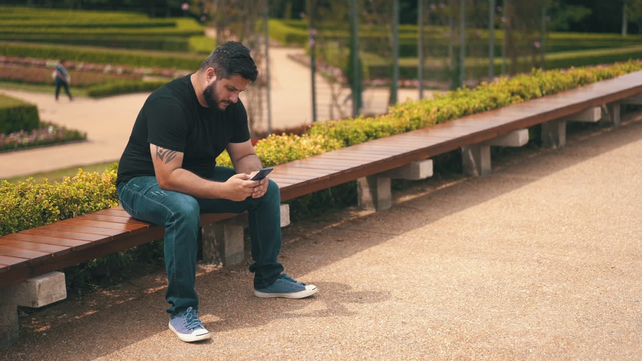 joven preocupado enviando mensajes de texto en su teléfono celular mientras estaba sentado en un banco largo en el parque al aire libre
