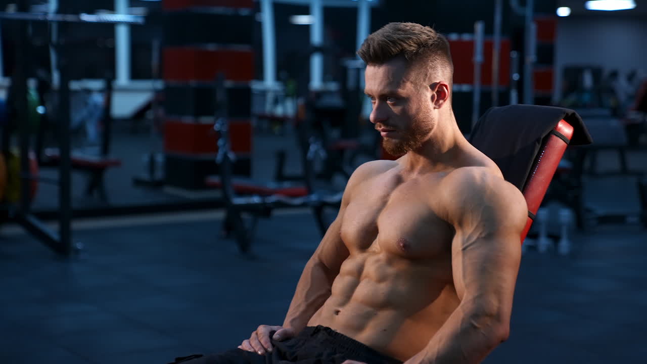 Strong handsome fitness trainer sits on sport chair and rests after training muscles. Naked torso with perfect muscles.