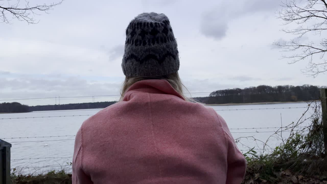 mujer rubia de moda con abrigo rosa y gorro disfrutando de una hermosa vista del lago natural y el paisaje natural