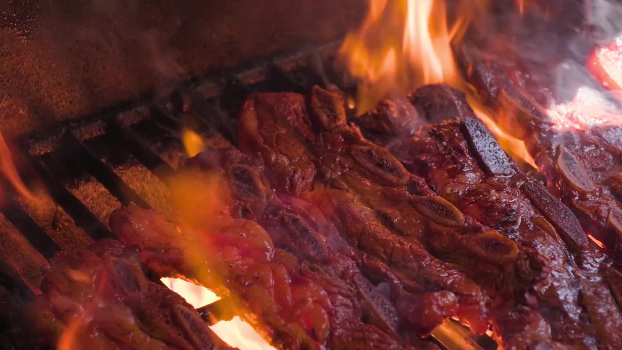 Imagens em close-up de churrasco, mostrando em câmera lenta chamas altas assando costelas bovinas durante o preparo na grelha quente