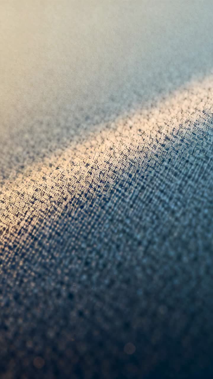 Striscia di luce calda in movimento che attraversa un tessuto intessuto blu-grigio mentre l'obiettivo si sposta in macro, mostrando i fili