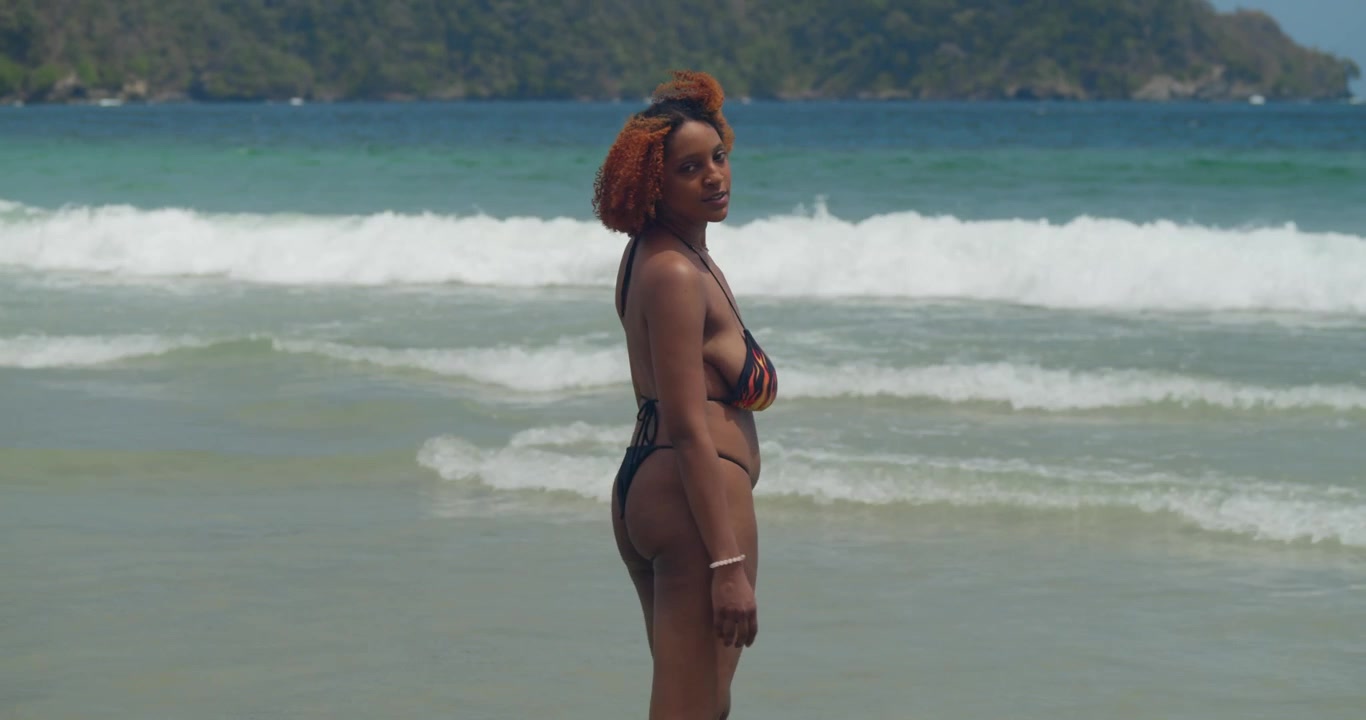 Una splendida donna dai capelli rossi in un elegante bikini nero si erge su una spiaggia caraibica incontaminata, sorridendo calorosamente alla macchina fotografica.