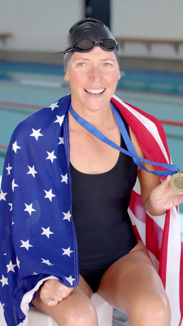 Vertical video: Smiling female swimmer holding gold medal and wearing American flag in pool area