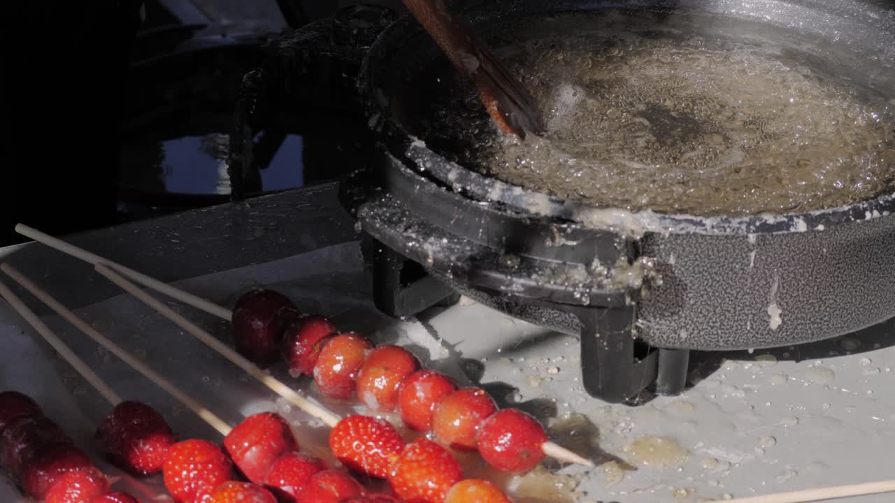 Making fruits in boiling caramel close-up. Chinese winter sweet dessert Tanghulu street food.Berries, grapes, tangerine and others fruit caramelized in sugar. Asian street market with traditional food.. High quality 4k footage