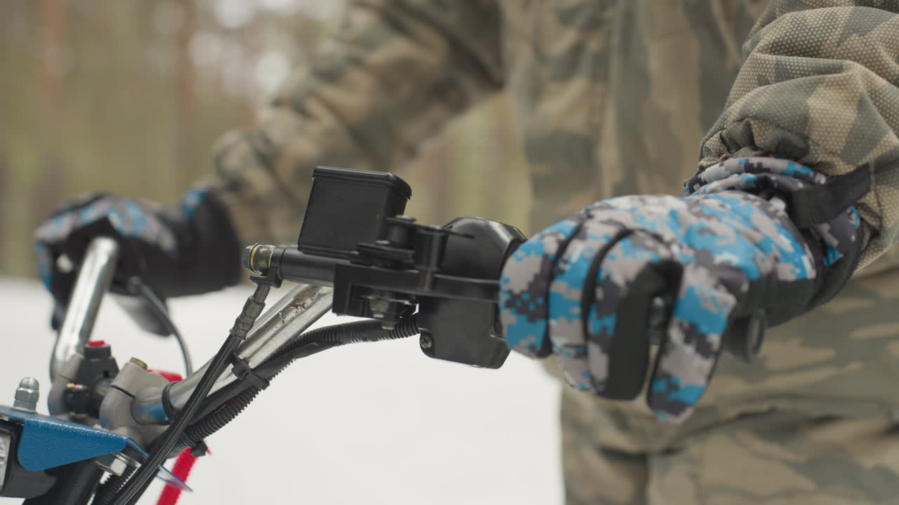 Mani guantate sul manubrio dell'ATV, giacca mimetica in foresta innevata, regolazione dell'acceleratore e del supporto della telecamera, primo piano di guanti invernali isolati che dimostrano presa sicura e controllo preciso durante un percorso estremo.