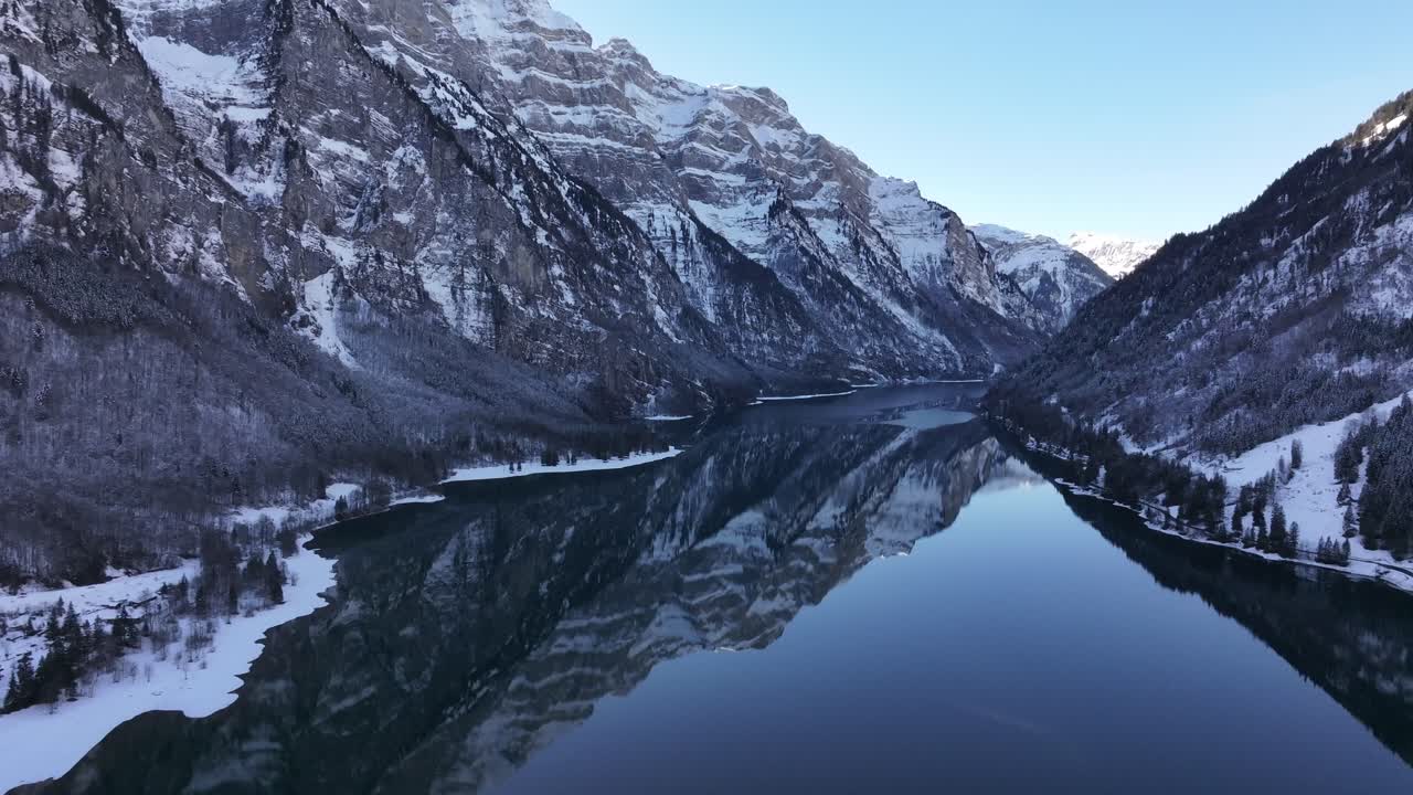 Drone flight over Klöntalersee lake in Switzerland, capturing the stunning reflection of the surrounding mountains on its calm waters.