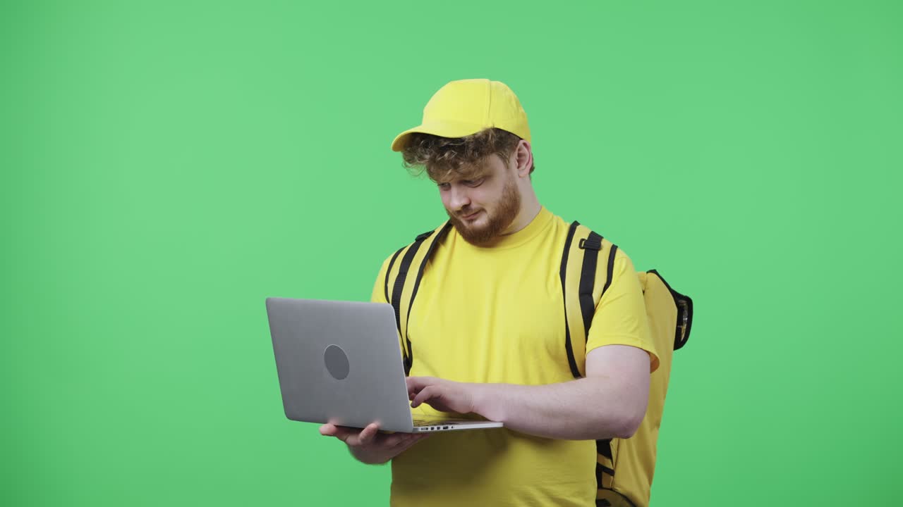 retrato de hombres jóvenes escribiendo en el teclado de la computadora portátil. hombre pelirrojo en uniforme de mensajero de entrega amarillo con bolsa térmica posando en la pantalla verde en el estudio. primer plano. cámara lenta lista 59.97fps