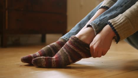 woman putting on woolen, striped socks while sitting on wooden floor in apartment