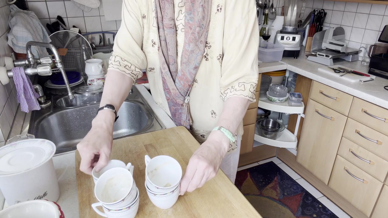 Woman begins sorting cups, plates and kitchen items from the sink onto a countertop before washing. Static Medium Shot.