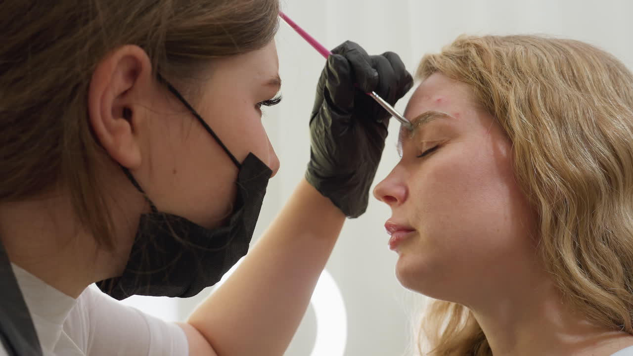 Beautician wearing black gloves carefully applies cream to client eyebrow using pink-handled brush in clean, well lit room with blurred ring light in background