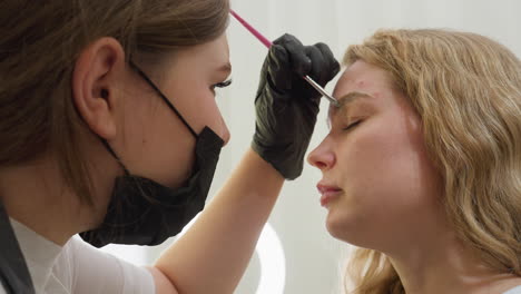 beautician wearing black gloves carefully applies cream to client eyebrow using pink-handled brush in clean, well lit room with blurred ring light in background