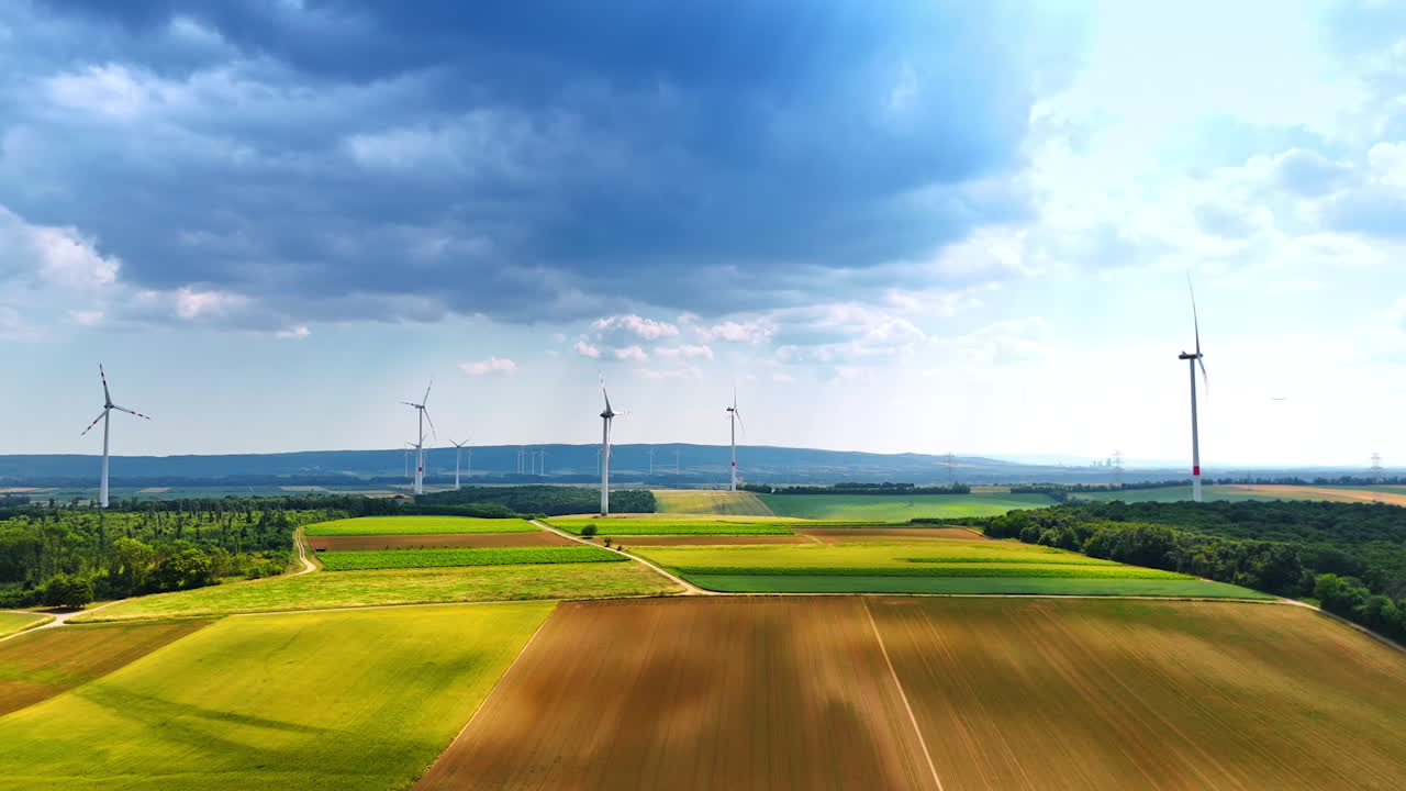 Wind turbines on a lush farm landscape. Vast fields of green and brown stretch beneath dark clouds with wind turbines generating clean energy in the distance