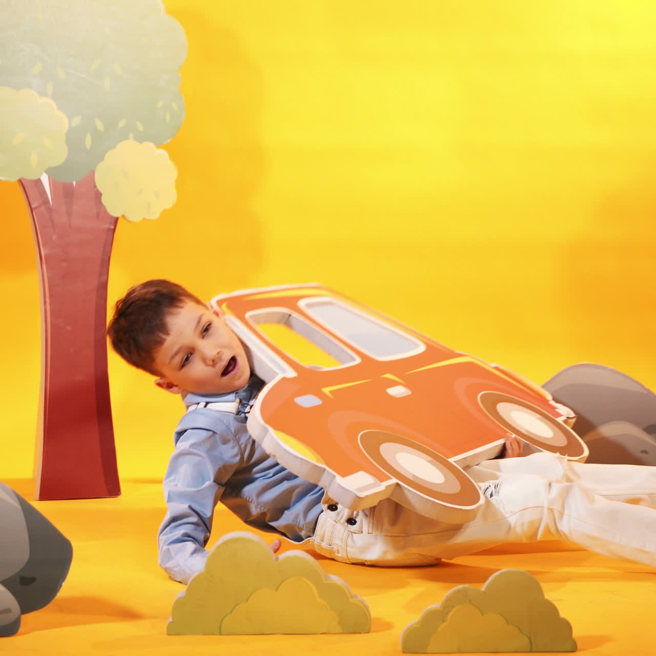 Cheerful little boy plays in the studio with toyful scenery on the yellow background. Happy kid with toyful carton car in his hands.