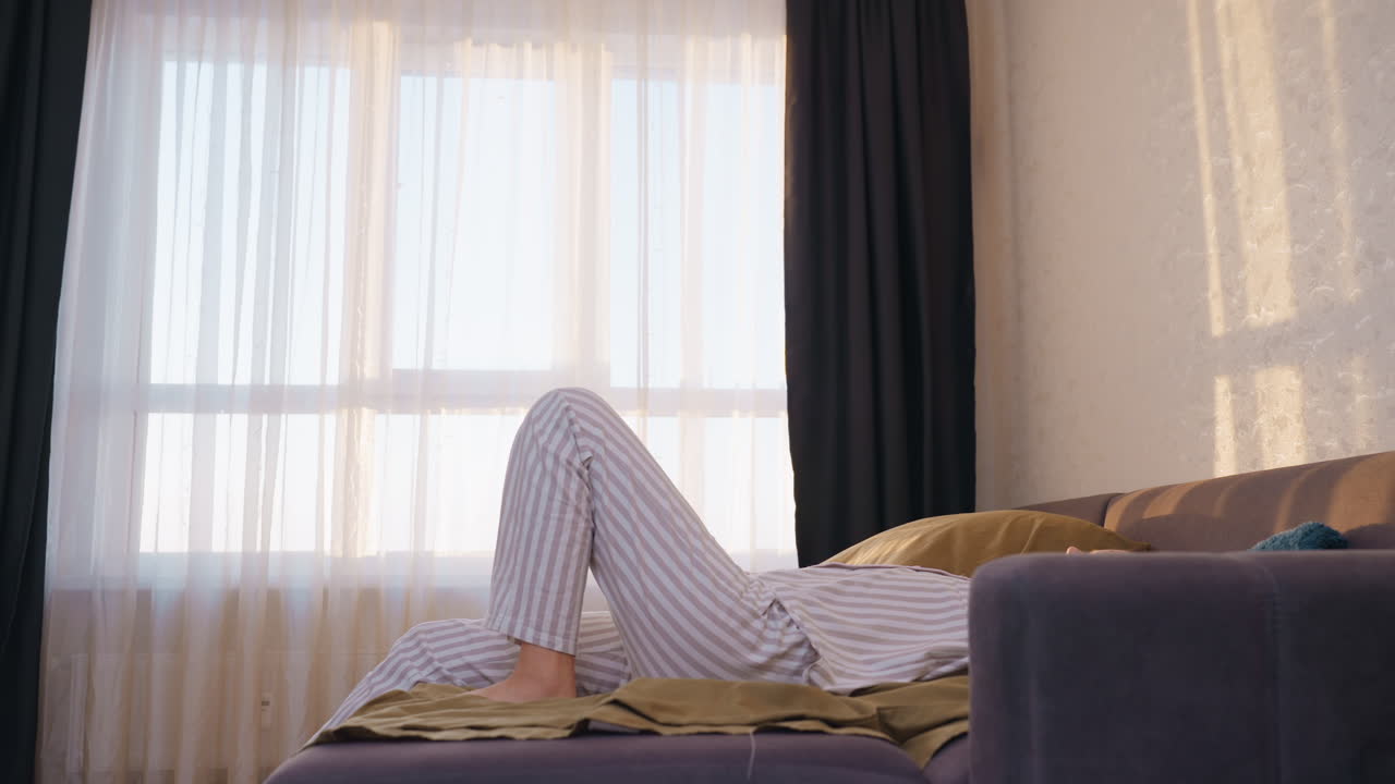 Young Woman Lounging Comfortably At Home, Casual Scene Of Woman Resting On Sofa Bed During Daylight, Relaxed Young Female Reclining On Couch In Striped Pajamas Through Softly Lit Window