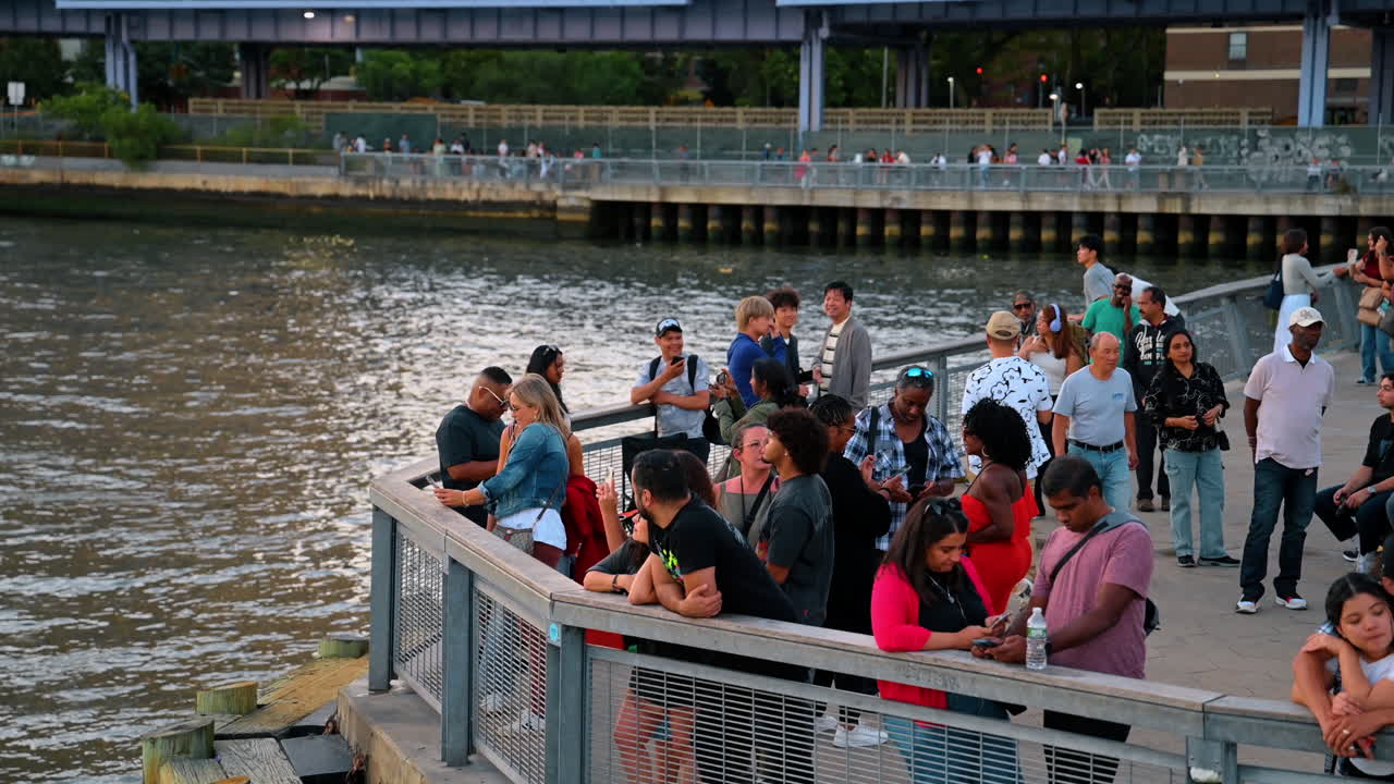 New York, États-Unis, 2 août 2025 : Groupe de personnes diverses rassemblées sur une jetée au bord de l'eau au crépuscule. Foule de gens debout et discutant sur une jetée en bois au bord du fleuve en soirée à New York.