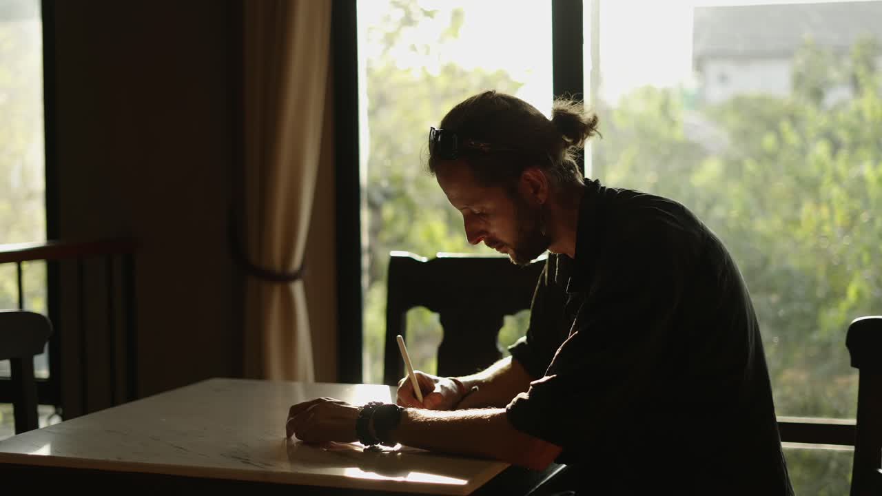 Man writing or drawing at a table by a window