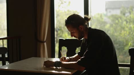 man writing or drawing at a table by a window