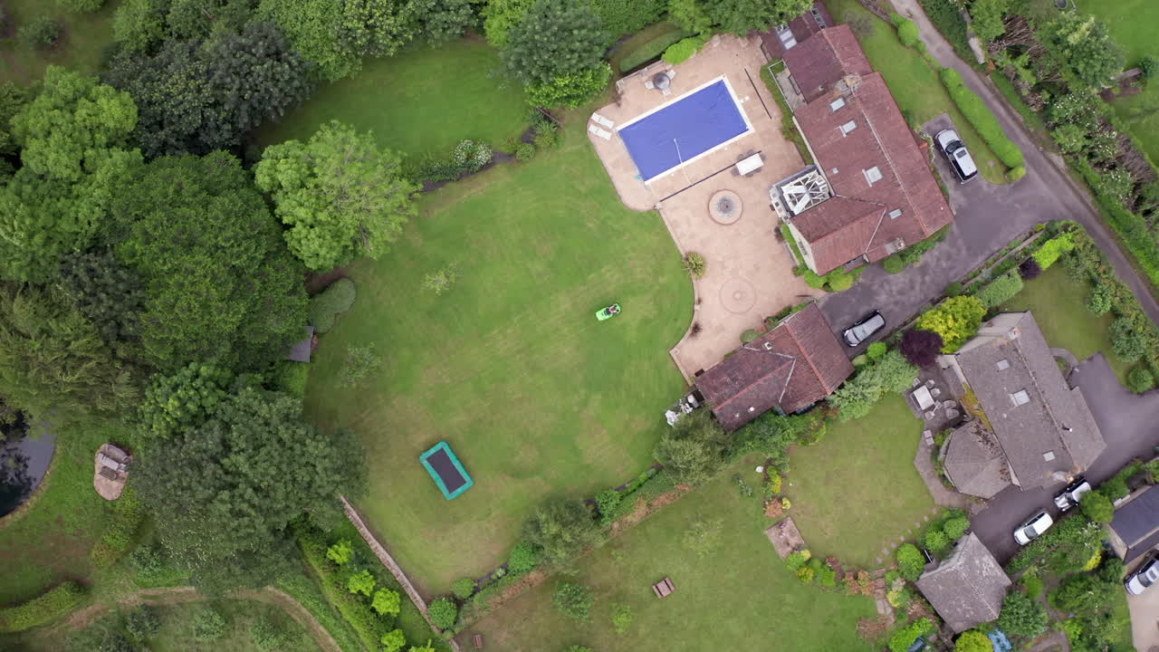 Falling Aerial Shot of Man Mowing Lawn of Home on Summer’s Day using Ride On Lawnmower from Birds Eye View Perspective