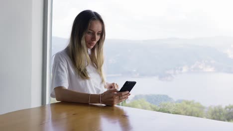 Girl-surfing-internet-at-her-phone-while-sitting-with-the-panoramic-window-landscape-view