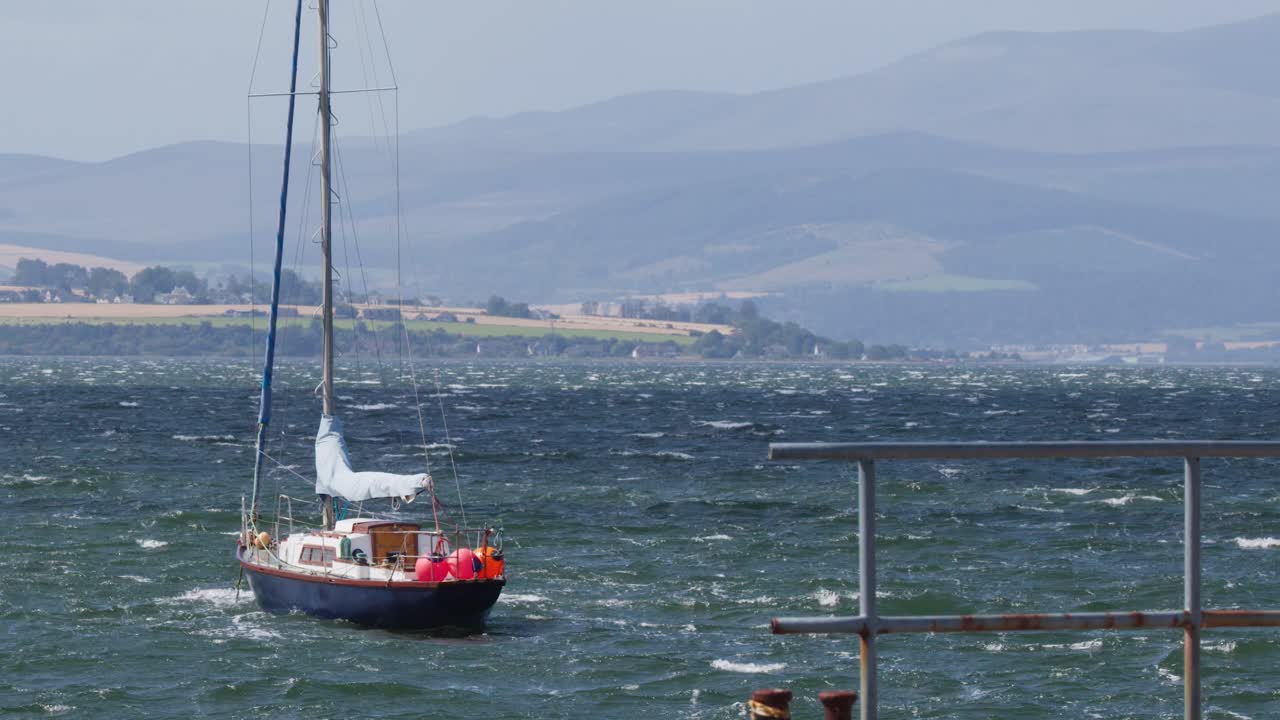 Red sailing boat moves toward marina in choppy waters, distant Scottish Highlands under daylight
