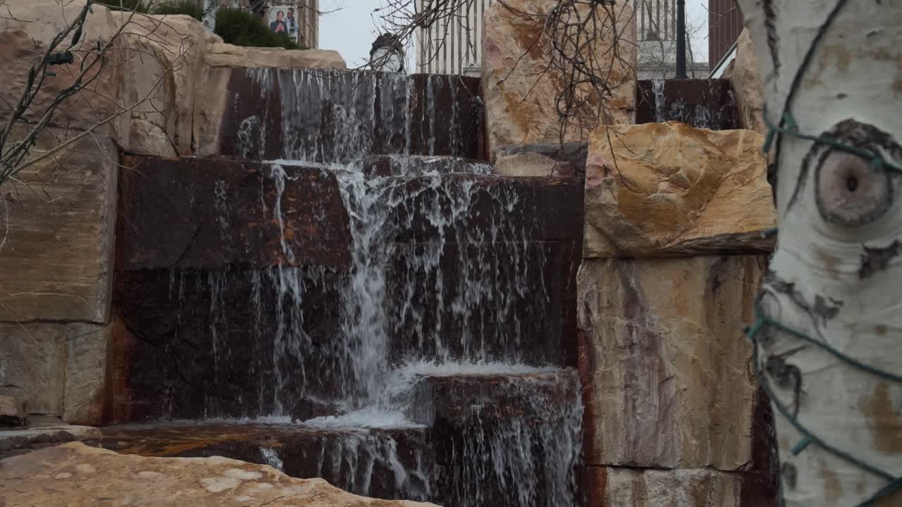 Camera pans up from greenery to reveal cascading stone waterfall in public park