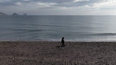 toma aérea de seguimiento de una mujer paseando a un perro pequeño en una playa vacía, día nublado