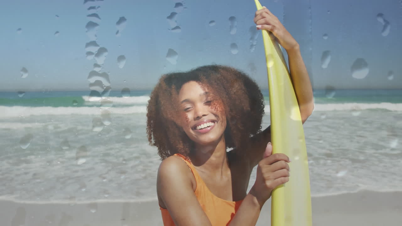 Animation of water droplets over smiling african american woman at beach with surfboard over sea