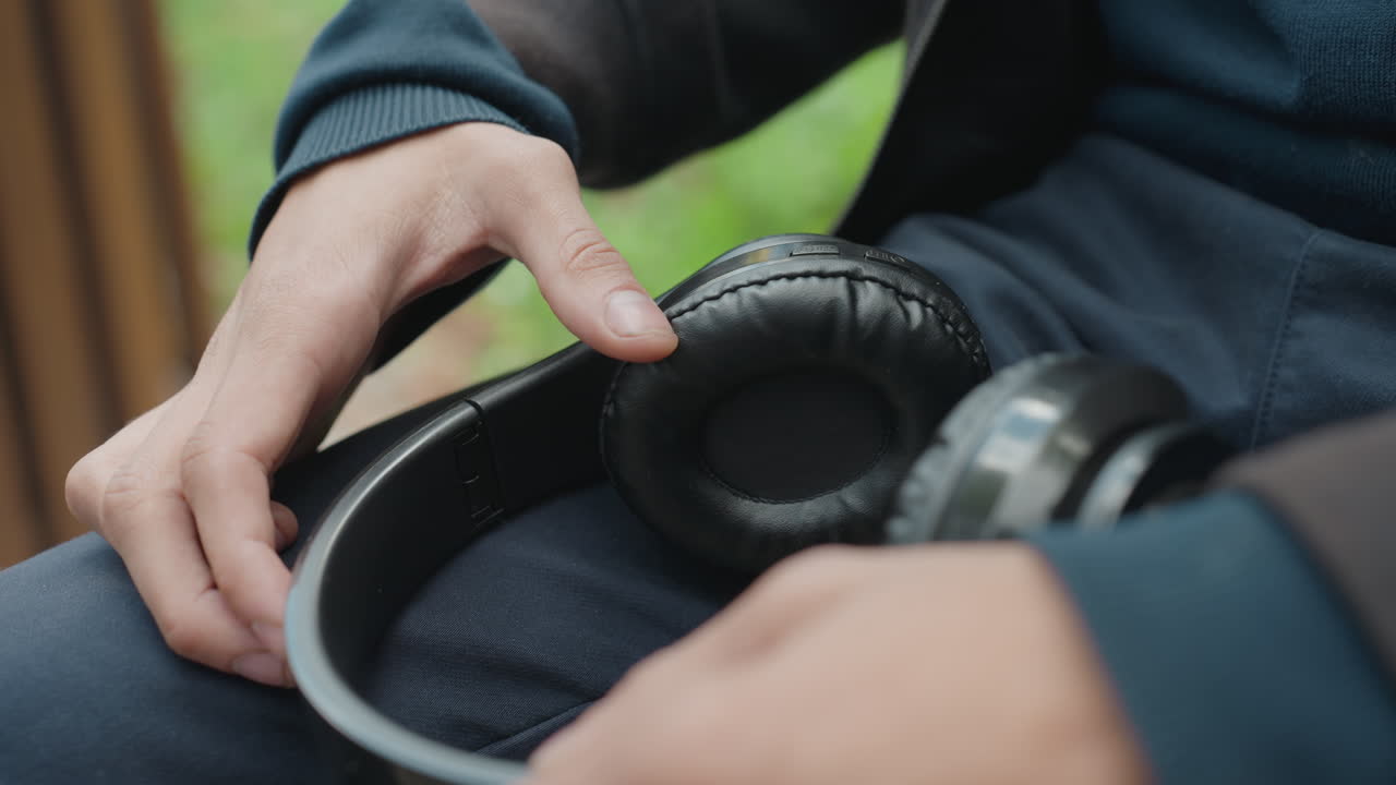 man inspecting worn leather headphones, on lap near window, fingers tracing torn earcup and frayed cushion, natural light, calm reflective mood, blurred garden background, denim pants and navy jacket