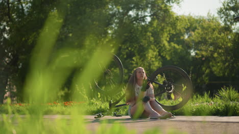 young lady short jean and blonde hair seated near her upside-down bike answering a phone call, surrounded by vibrant greenery and trees in outdoor setting