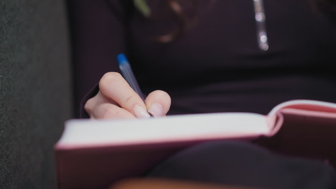 Close up view of woman's hand with manicured nails holding blue pen while writing in pink notebook on lap, captured against dark clothing in relaxed indoor setting with focus on calm concentration