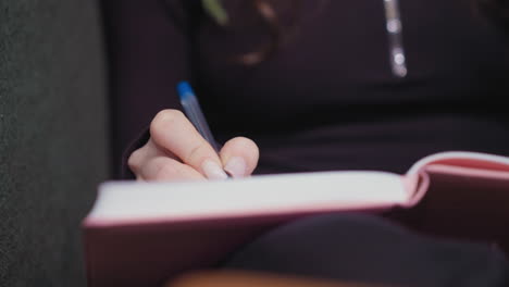 close up view of woman's hand with manicured nails holding blue pen while writing in pink notebook on lap, captured against dark clothing in relaxed indoor setting with focus on calm concentration