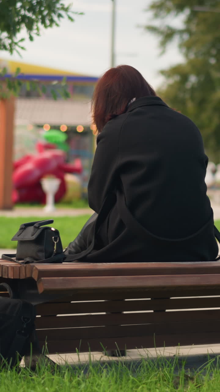 공원 벤치에 앉아있는 여성의 뒷면, 검은 코트와 부츠를 입고, 편안한 도시 공원 분위기에서 푸른 나무로 둘러싸여, 놀이터와 다른 공원 특징으로 흐릿한 배경