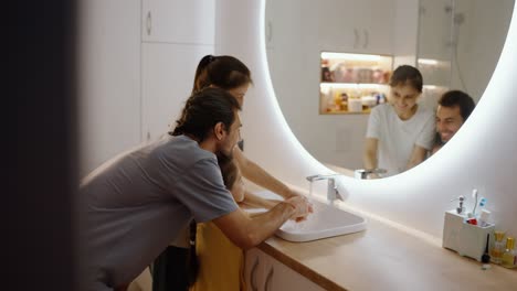 desde la parte de atrás, un hombre moreno con una camiseta gris junto con su esposa, una chica morena con una camisa blanca, se lavan las manos con su hija pequeña en un vestido amarillo en un fregadero moderno con un espejo en el baño
