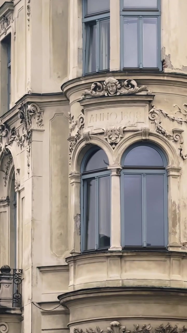 Primer plano vertical de la Casa Frisch en Liubliana, mostrando un ornamentado mirador esquinero histórico con ventanas arqueadas pareadas, un adorno facial esculpido y detalles decorativos de estuco.