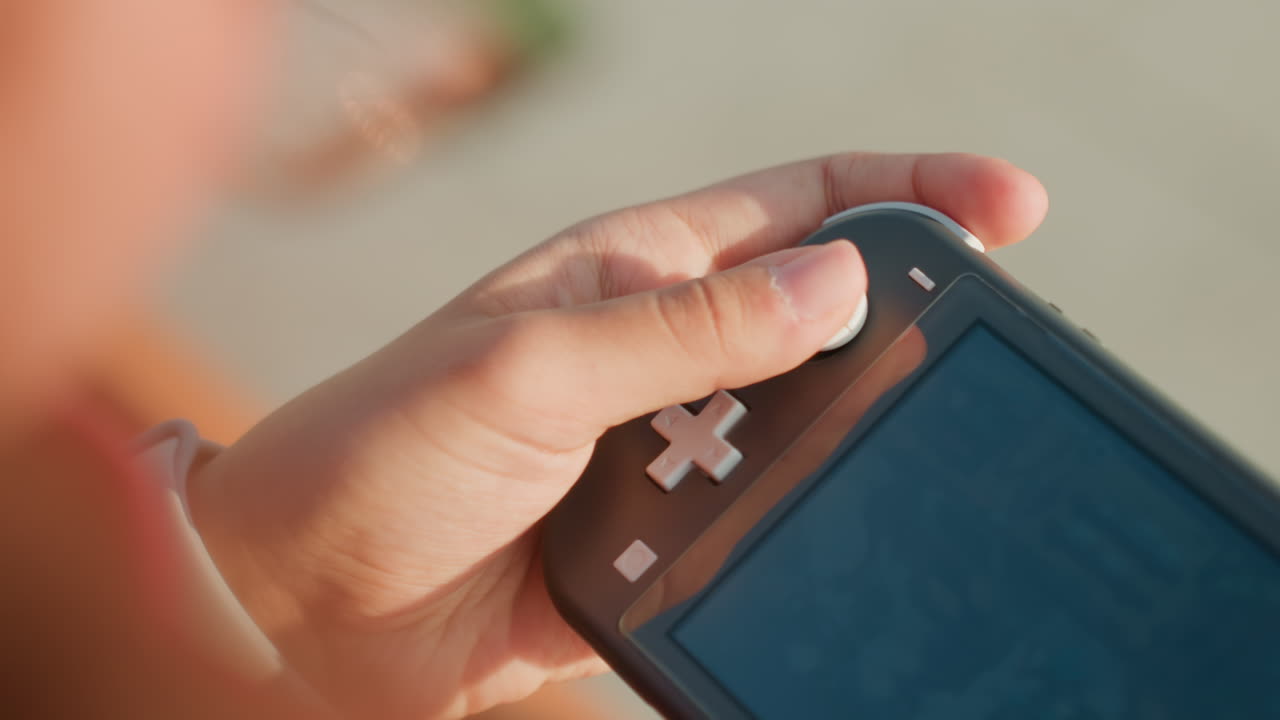 Close-up van kleine handen die een handheld console spelen met de duim op de D-pad, zonnige buitenomgeving, oprechte zomerse sfeer, geconcentreerde jonge speler oefent de bediening op een draagbaar apparaat, zachte bokeh en warme tinten