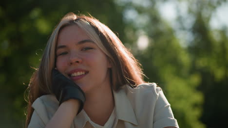 close-up of young woman resting her hand on her chin with a gentle, contemplative smile, sunlight softly illuminates her face, natural glow against a blurred background of vibrant green foliage