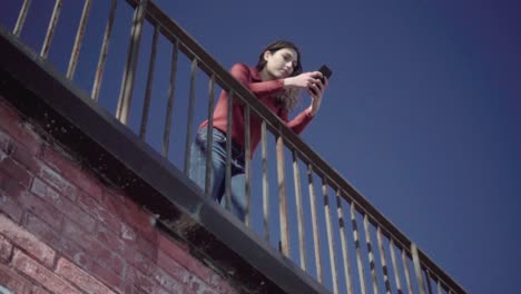 chica apoyada en una barandilla usando el teléfono inteligente en cámara lenta con cielo azul en el fondo