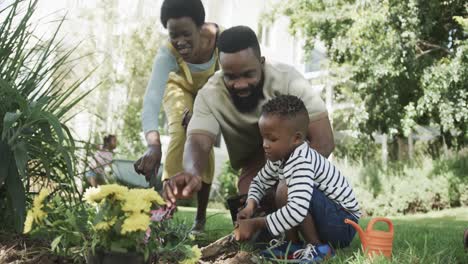 Feliz-Pareja-Afroamericana-Con-Su-Hijo-Plantando-Flores-En-Un-Jardín-Soleado,-Cámara-Lenta