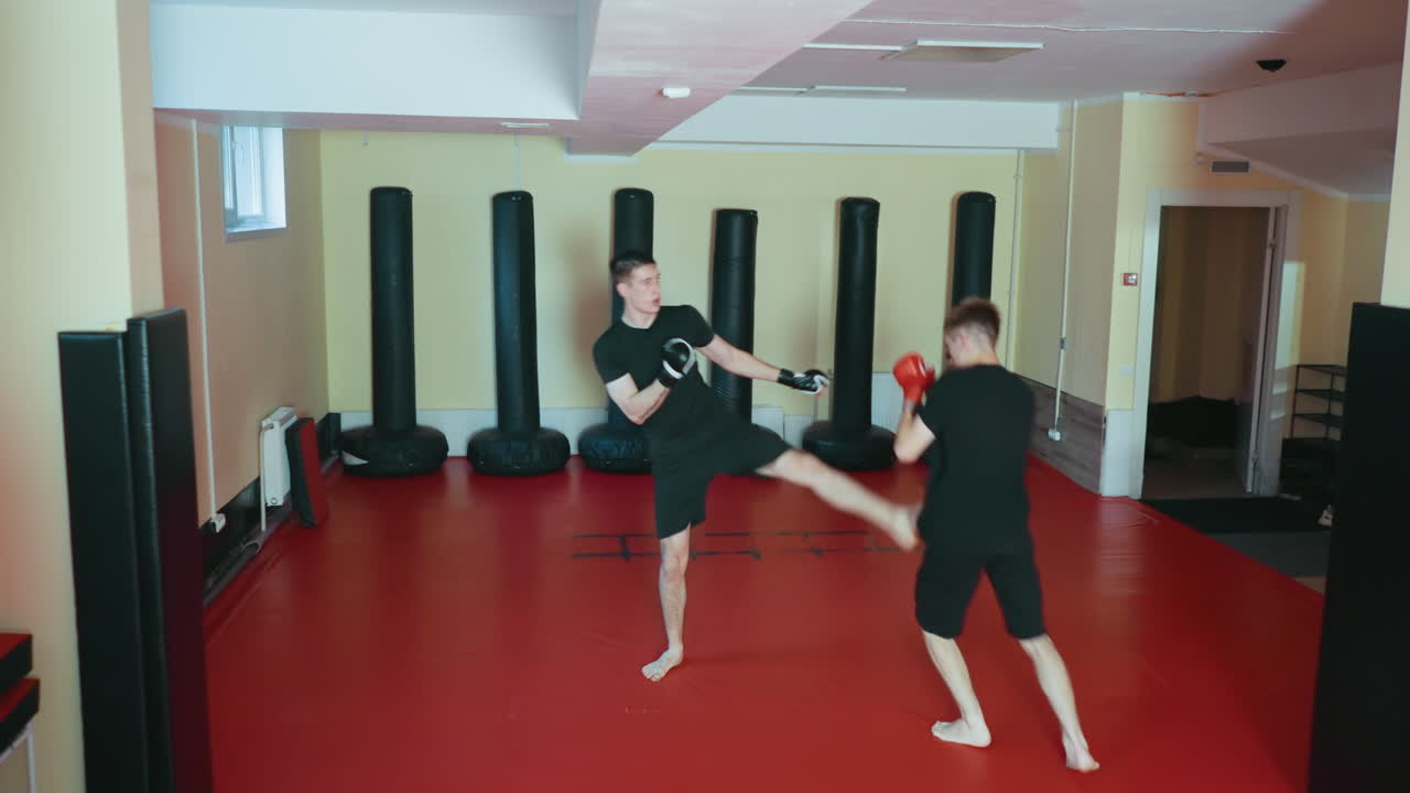 Atletas practicando sparring en un gimnasio de artes marciales sobre una colchoneta roja, con guantes, centrándose en el entrenamiento de combate, el juego de pies y la técnica, mostrando energía, equilibrio y coordinación durante la sesión de entrenamiento