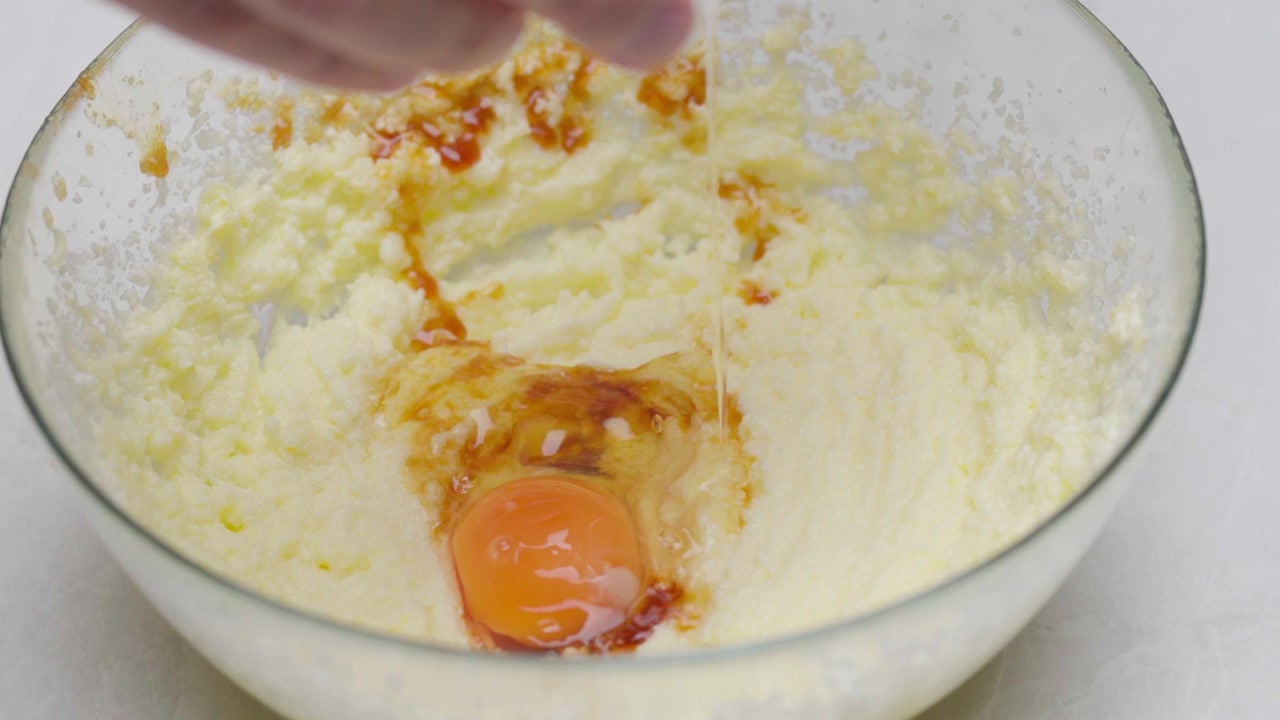 Close-up as raw egg is added to creamy mixture inside a glass bowl.