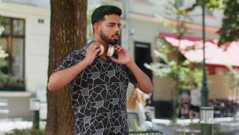happy young indian man putting on wireless headphones listening music on smartphone passes street